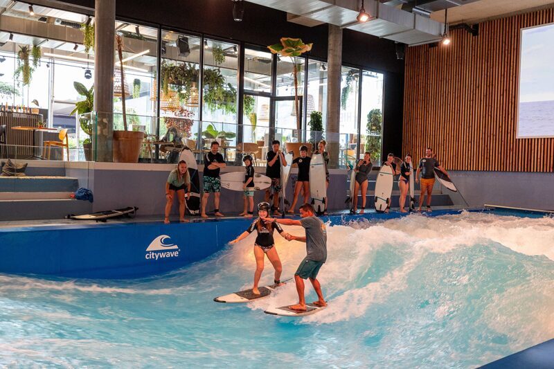 Zwei Personen surfen auf einer künstlichen, indoor Welle, einige Personen schauen zu